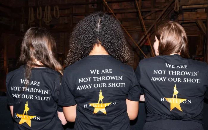 Backs of three students wearing Hamilton tee shirts that say "We are not throwin' away our shot!"