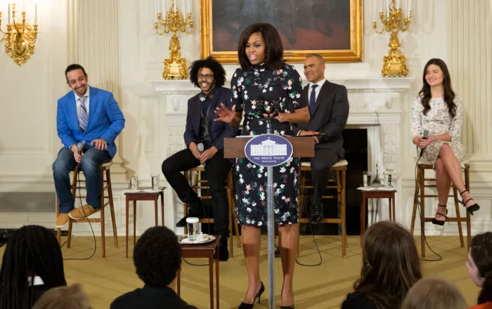 Michelle Obama speaking at White House podium with Lin-Minuel Miranda and other Hamilton cast members seated behind her